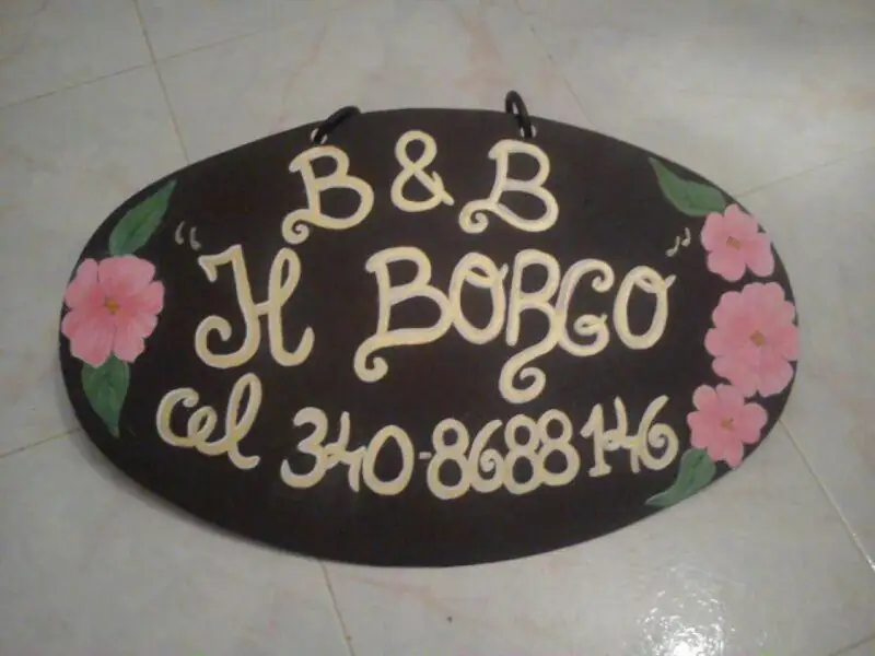 Insegna ovale dipinta a mano del B&B Il Borgo, con lettere chiare e fiori rosa su fondo scuro.