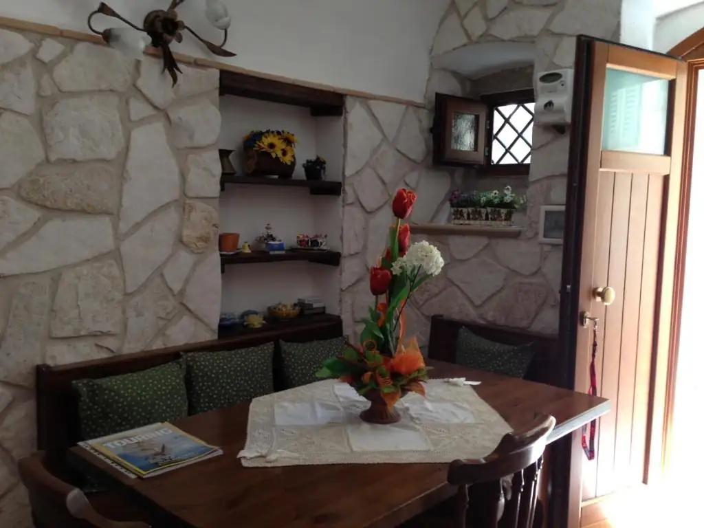 Soggiorno de Il Borgo 19: parete in pietra chiara, tavolo in legno con centrino e fiori, panca con cuscini verdi e mensole con oggetti in ceramica.
