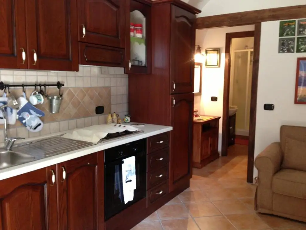 Cucina de Il Borgo 19 con mobili in legno scuro, lavello, forno incassato e rivestimento in piastrelle chiare; sul fondo il passaggio verso il bagno.