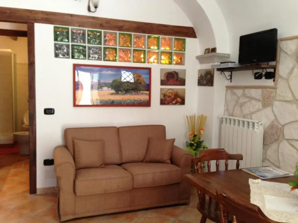 Divano letto beige nel soggiorno de Il Borgo 19, con quadri alle pareti, parete di vetrocemento colorato, televisore e pavimento in cotto.