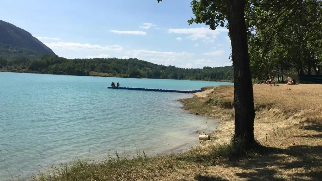Riva del lago di Castel San Vincenzo con una passerella galleggiante che entra nell’acqua turchese, alberi e montagne.