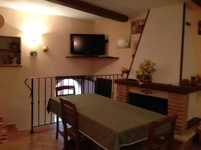 Zona pranzo de Il Borgo 30: tavolo apparecchiato con tovaglia verde, camino, televisore e la ringhiera della scala interna.