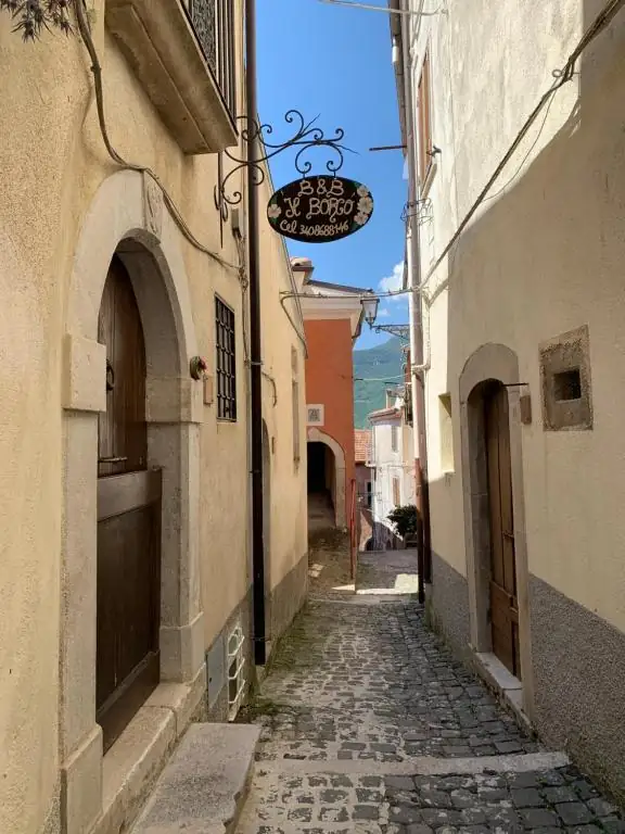 Vicolo lastricato del centro storico di Scapoli, con l’insegna in ferro battuto del B&B Il Borgo appesa fra due case in pietra e le montagne sullo sfondo.